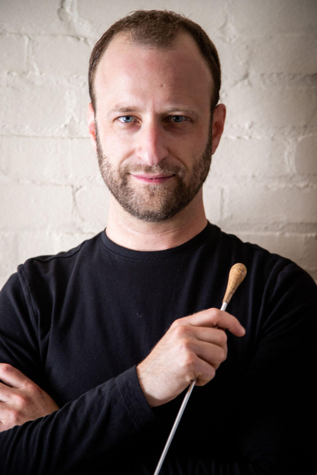 Dr. Matthew Mainella looks at the camera with a small, relaxed smile. The lighting is dramatic, highlighting the left side of his face and casting a shadow on the right side. His conducting baton is held loosely in one hand across his chest.