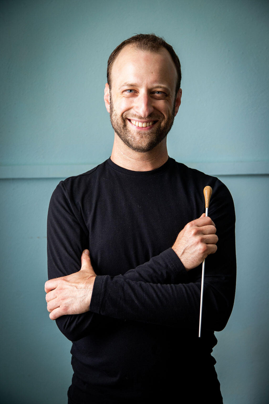 Photo by Wendy Redfern Dr. Matthew Mainella stands with his arms confidently crossed, a conductor baton held upside down in one hand. He is wearing all black, casual clothes and smiling broadly at the camera. The light blue-green background perfectly matches his eyes.