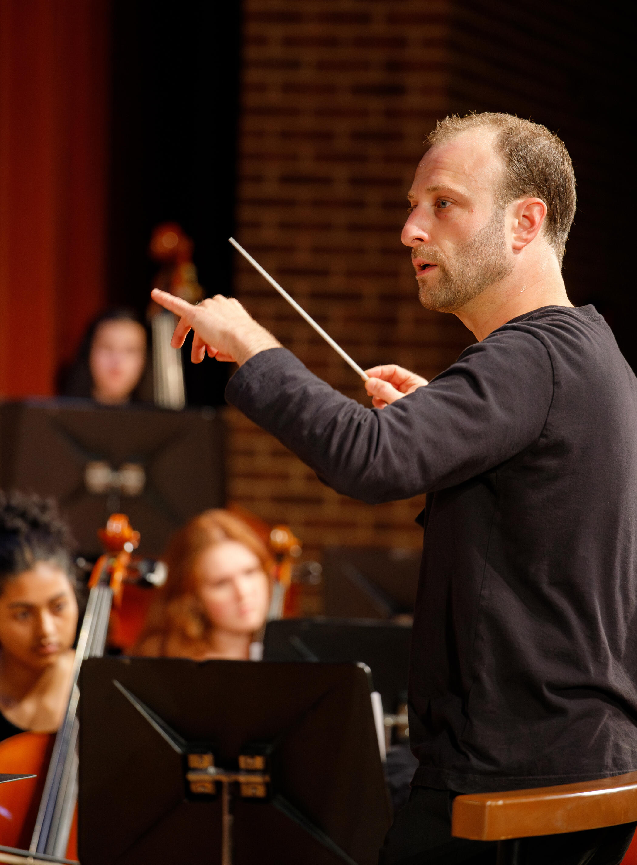 Dr. Matthew Mainella stands in front of a polished wooden rail, hands lifted, with a white baton in one. His mouth is slightly open, and his focus is off to the left as he directs a youth orchestra. Blurred in the background are young orchestra members.