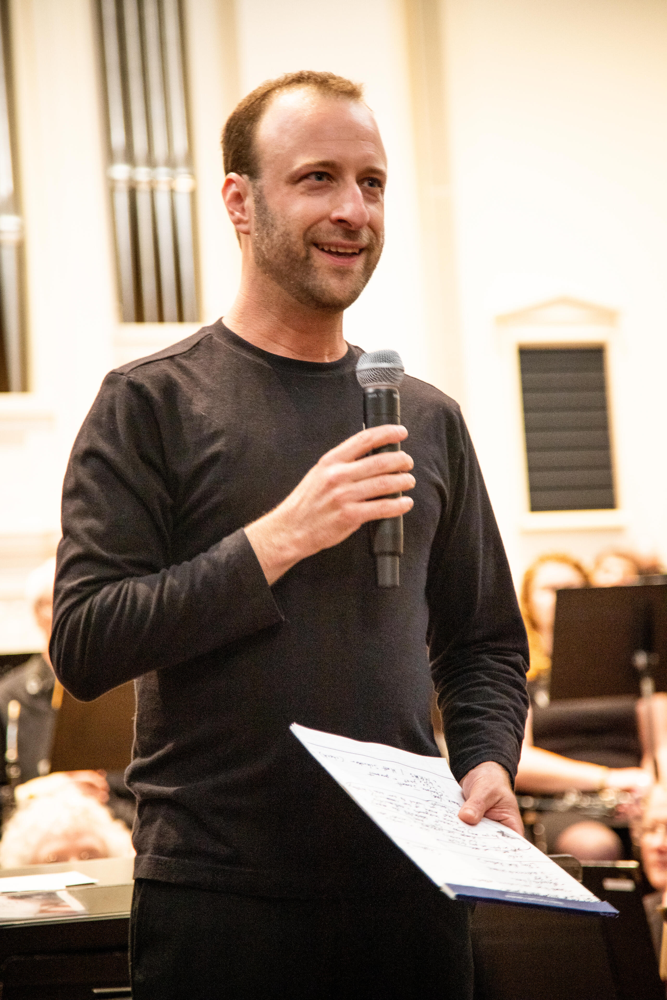 Photo by Wendy Redfern Dr. Matthew Mainella stands in front of an orchestra with a pipe organ blurred in the background. He holds a microphone and notepad, and he is smiling as he addresses the audience. He wears casual concert black.
