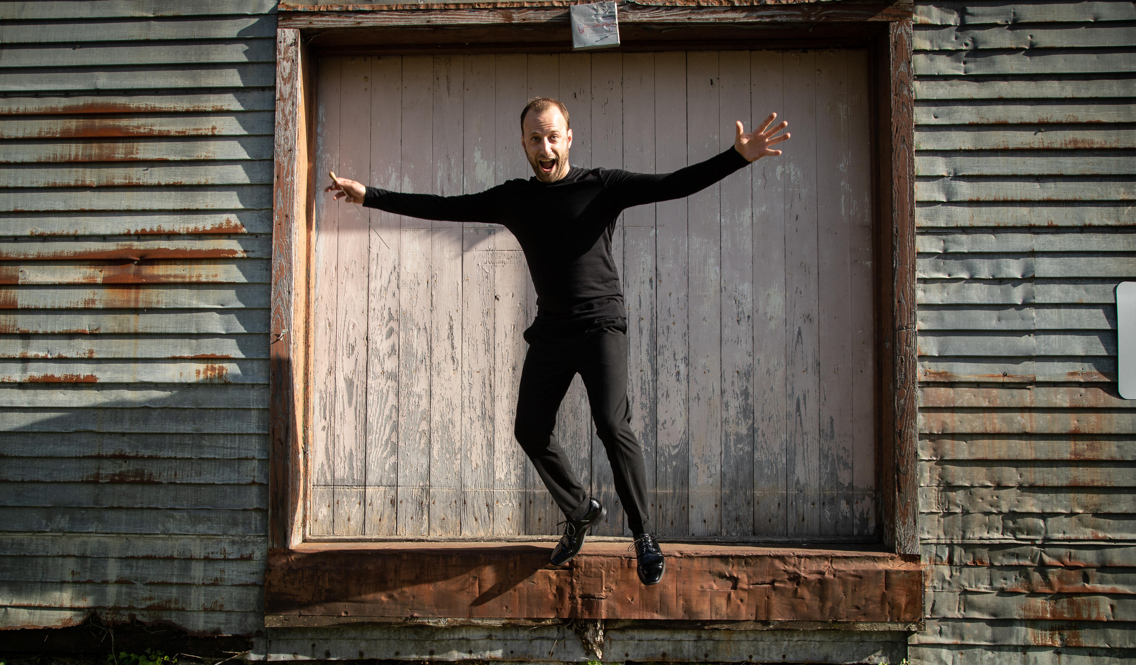 Photo by Wendy Redfern Dr. Matthew Mainella playfully jumps off a ledge in front of an old warehouse door. His arms are wide and his mouth is open in a funny expression, but he's wearing concert blacks and polished shoes, baton in hand.