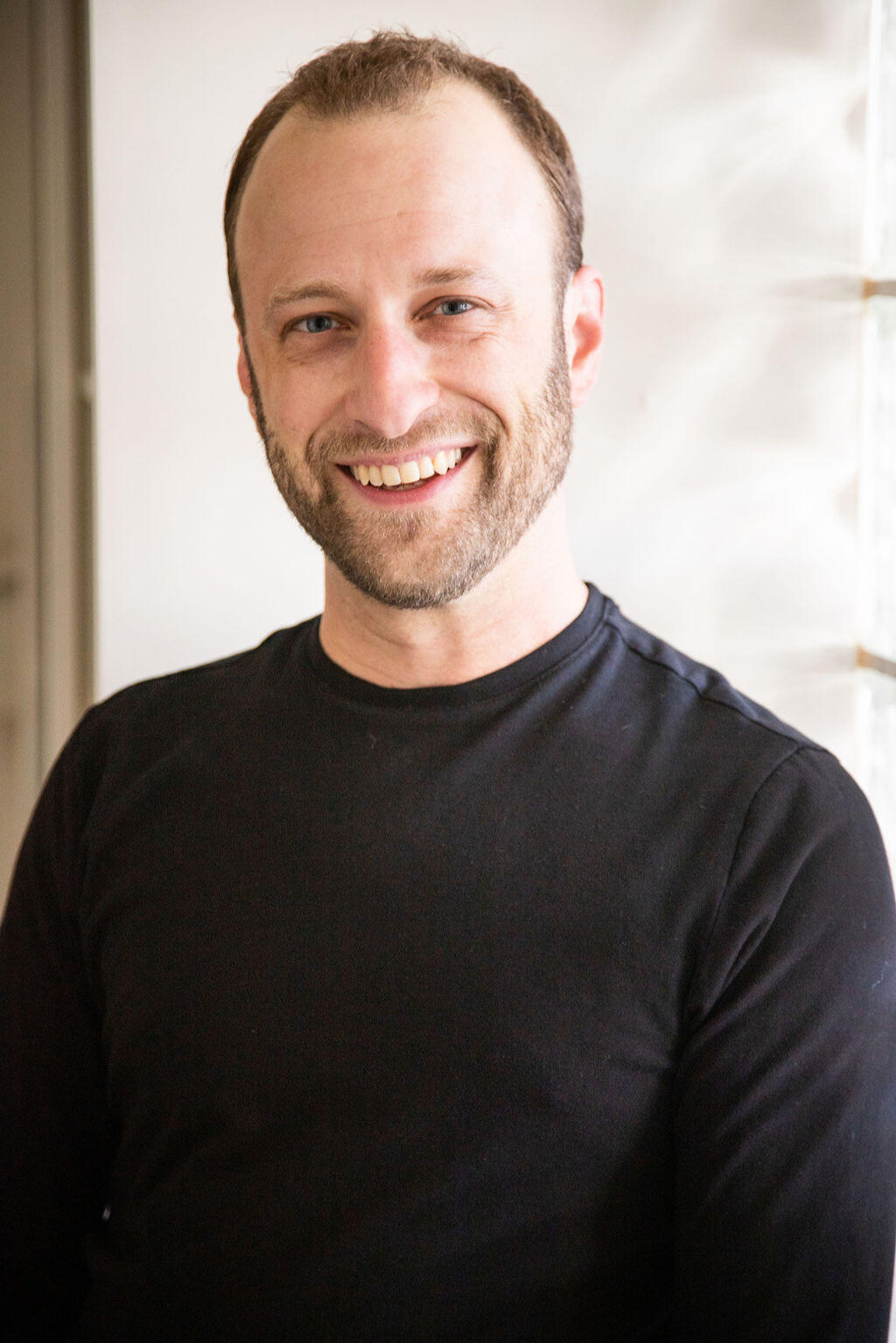 Dr. Matthew Mainella smiles at the camera, his head tilted to the side, his eyes focused on the viewer. He wears casual concert black and is leaning in front of a white-washed wall.